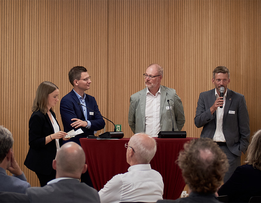 Auftaktveranstaltung MORO Fläche Personen sprechen bei einer Veranstaltung vor Publikum über das Modellvorhaben MORO Fläche